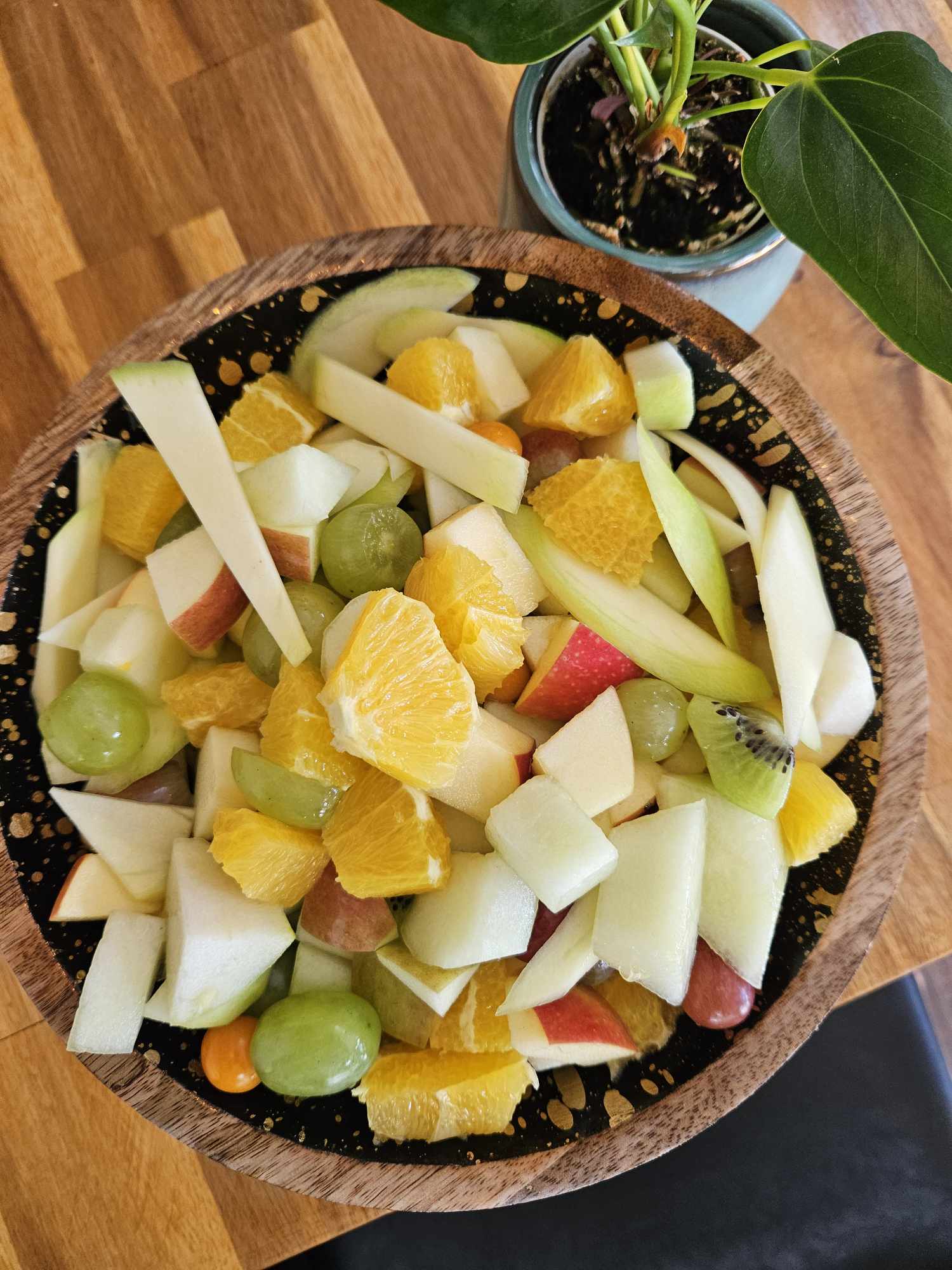 Bunter Obstsalat in Holzschale auf Holztisch; Pflanzenblatt rechts oben sichtbar.